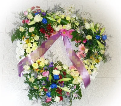Corona fúnebre de flores variadas con rosas blancas y azules, claveles y gerberas, con cinta lila